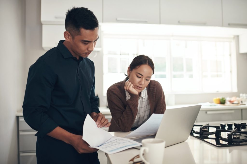 Concerned couple reviewing paperwork and using a laptop in their kitchen after receiving a denial for their fire damage insurance claim.